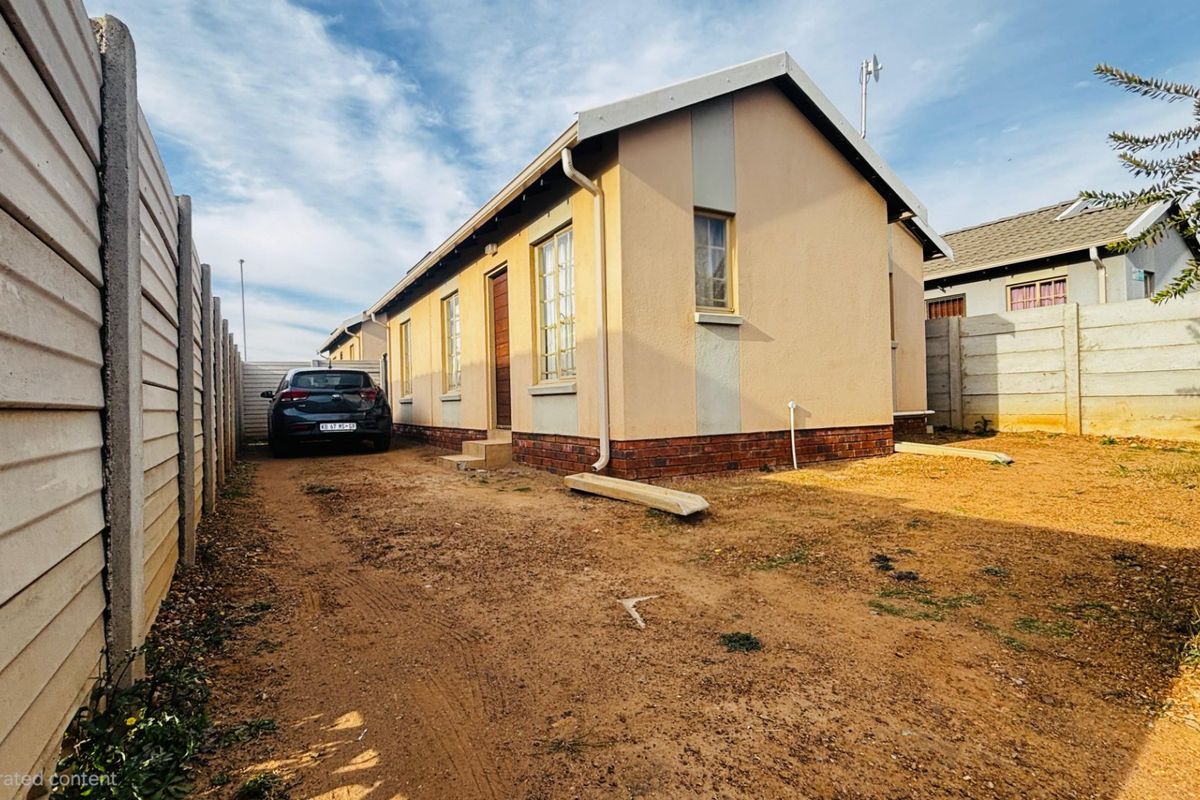 Rear view of the property featuring a precast concrete boundary wall, brick plinth detailing, and an unpaved driveway area.