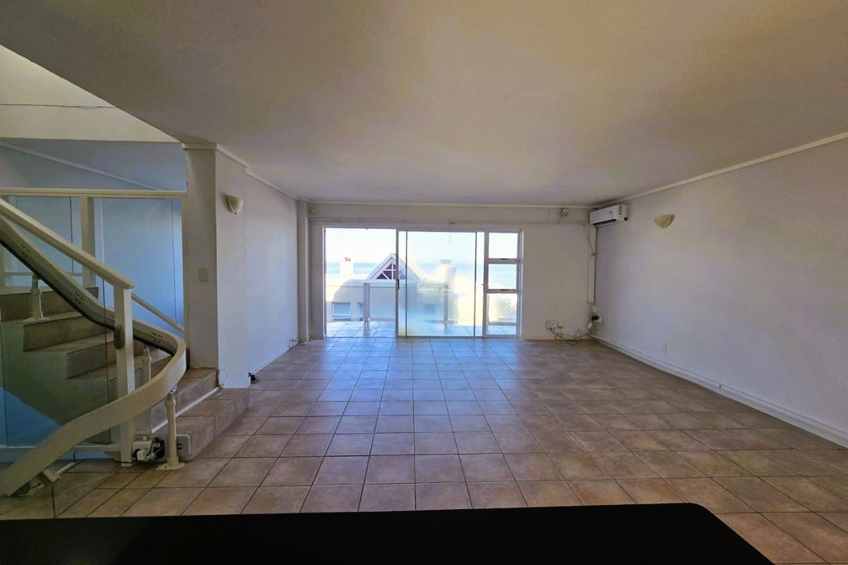 Spacious open-plan living area featuring tiled flooring, a curved staircase with glass balustrade, and large sliding doors leading to an outdoor balcony.