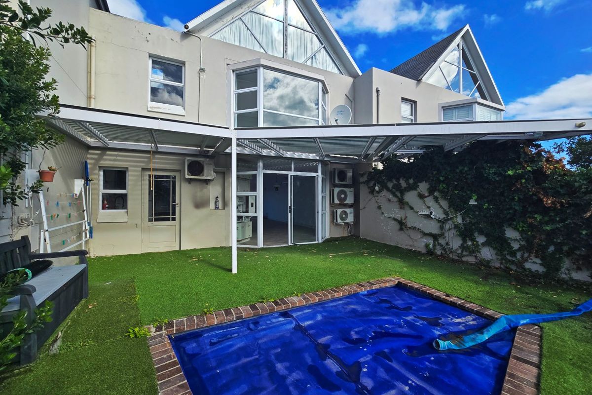 Rear exterior view of a townhouse featuring a swimming pool with a cover, artificial lawn, covered patio area, and distinctive gabled architectural windows.