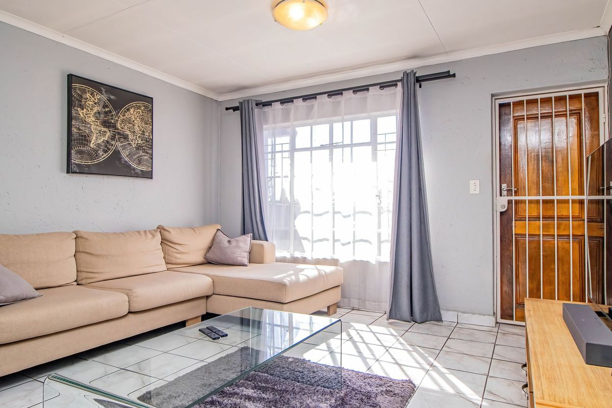 A living room featuring tiled flooring, a timber entrance door with a security gate, and natural light through a window.