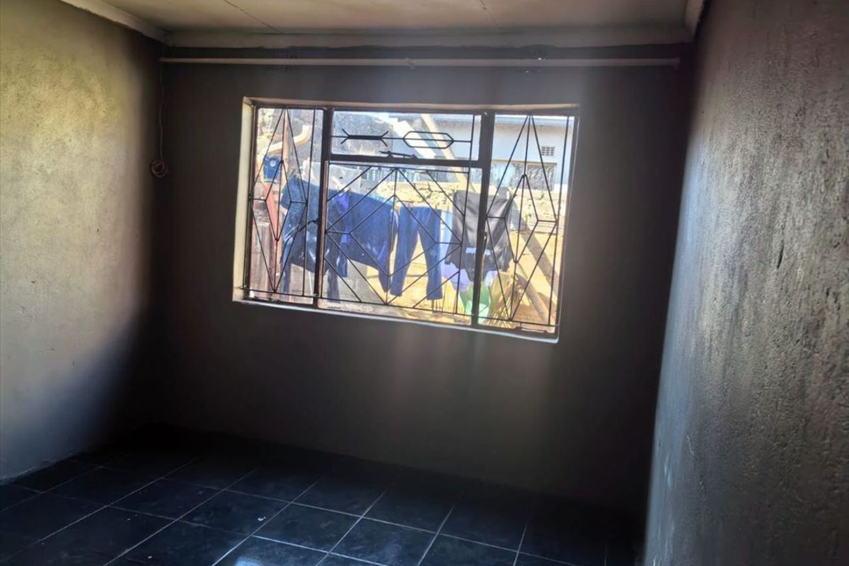 An empty bedroom featuring dark tiled flooring and security burglar bars on the window, requiring finishing touches to the wall surfaces.