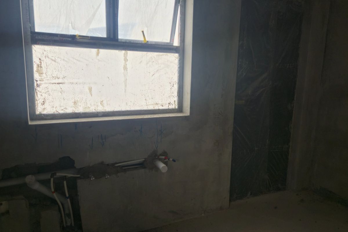 An unfinished kitchen space in shell condition featuring exposed plumbing and an installed aluminium window frame, ready for final fit-out and finishes.