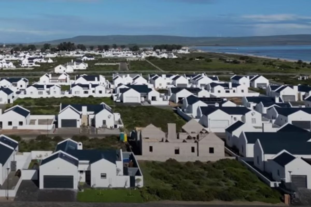 Aerial view of a coastal residential estate featuring uniform white houses with dark roofs, including a property currently under construction.