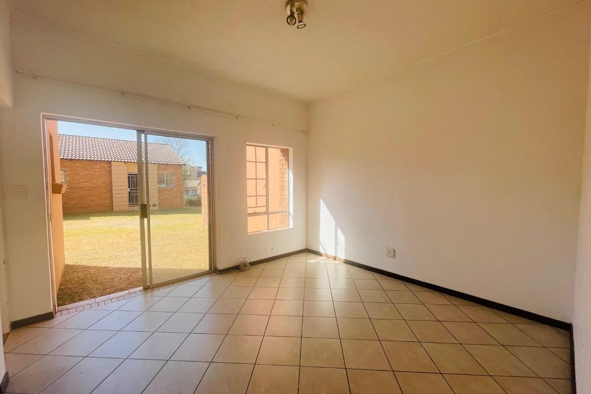 An empty, sunlit living room featuring neutral tiled flooring and a sliding glass door providing direct access to the outdoor garden area.