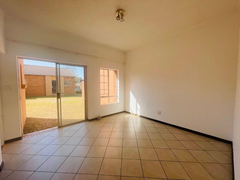 An empty, sunlit living room featuring neutral tiled flooring and a sliding glass door providing direct access to the outdoor garden area.