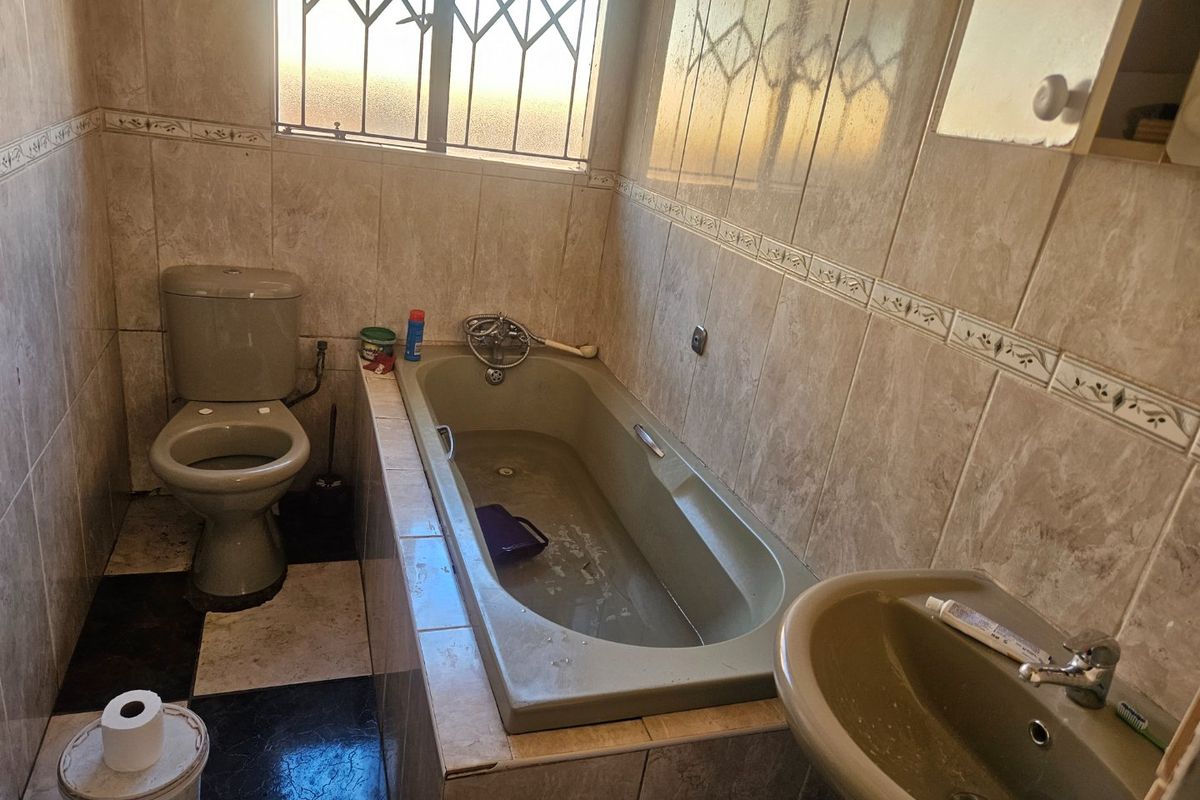 A dated bathroom featuring a built-in bathtub, pedestal washbasin, and toilet, finished with neutral wall tiles and security bars on the window.