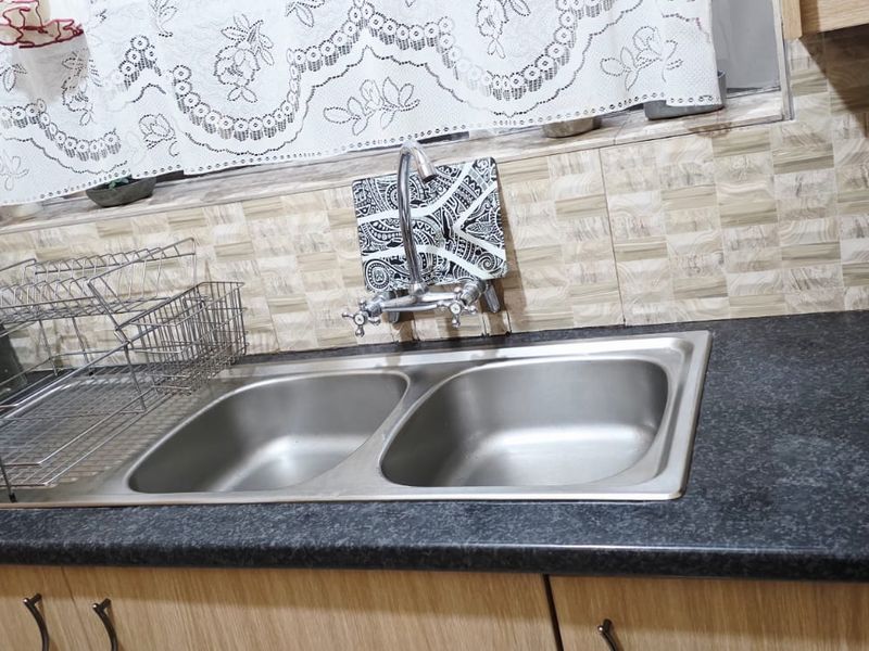 A functional kitchen area featuring a double bowl stainless steel sink, chrome mixer tap, dark laminate countertop, and tiled splashback with built-in cabinetry.