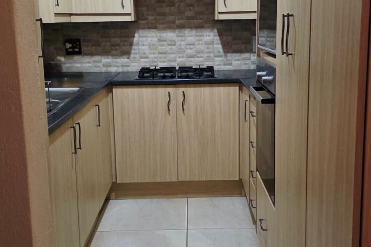 A compact, well-maintained kitchen featuring light wood-finish cabinetry, stone countertops, a gas hob, built-in oven, and neutral ceramic floor tiling.