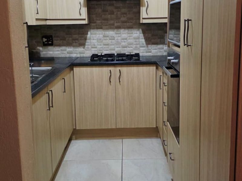 A compact, well-maintained kitchen featuring light wood-finish cabinetry, stone countertops, a gas hob, built-in oven, and neutral ceramic floor tiling.