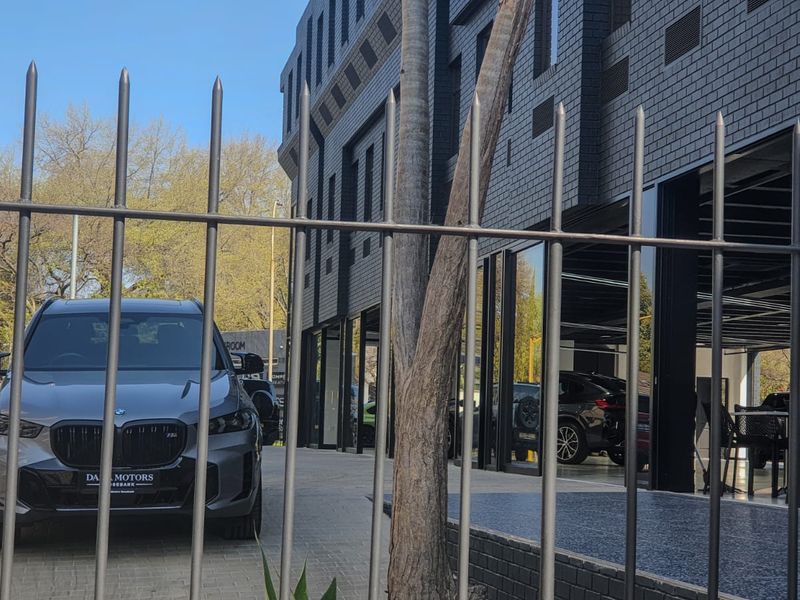 Modern commercial building facade featuring dark brickwork, extensive glass frontage, and a paved driveway, secured by metal perimeter fencing.