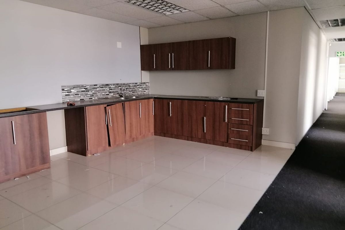 A vacant commercial kitchenette featuring dark wood-look cabinetry, tiled flooring, and a mosaic backsplash, currently requiring minor repairs to cabinet doors and hardware.