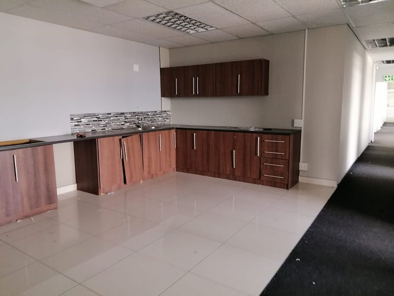 A vacant commercial kitchenette featuring dark wood-look cabinetry, tiled flooring, and a mosaic backsplash, currently requiring minor repairs to cabinet doors and hardware.
