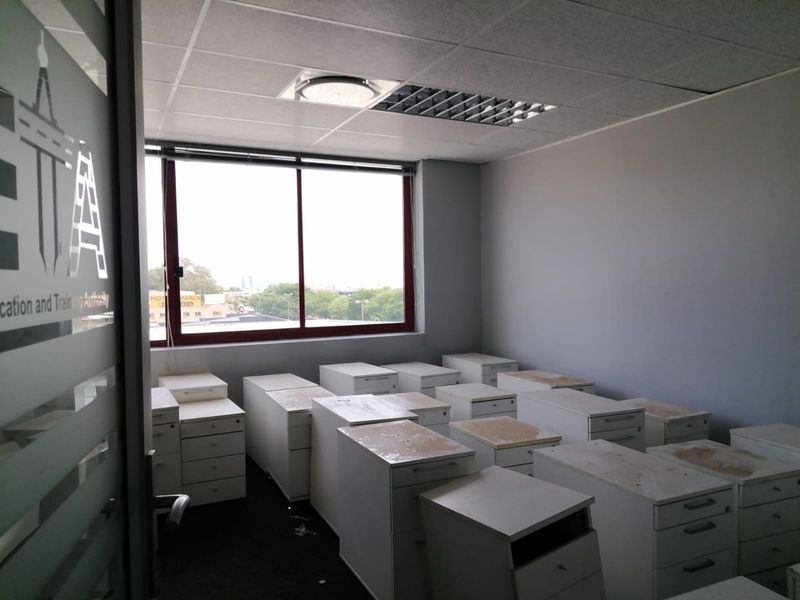 A commercial office space featuring a suspended ceiling, recessed lighting, and large windows, currently occupied by multiple storage pedestals.