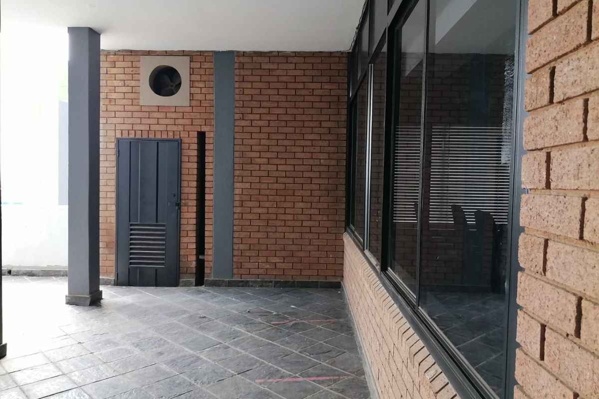 A covered outdoor patio area featuring exposed brick walls, tiled flooring, and large glass windows, providing a functional transition space for the commercial property.