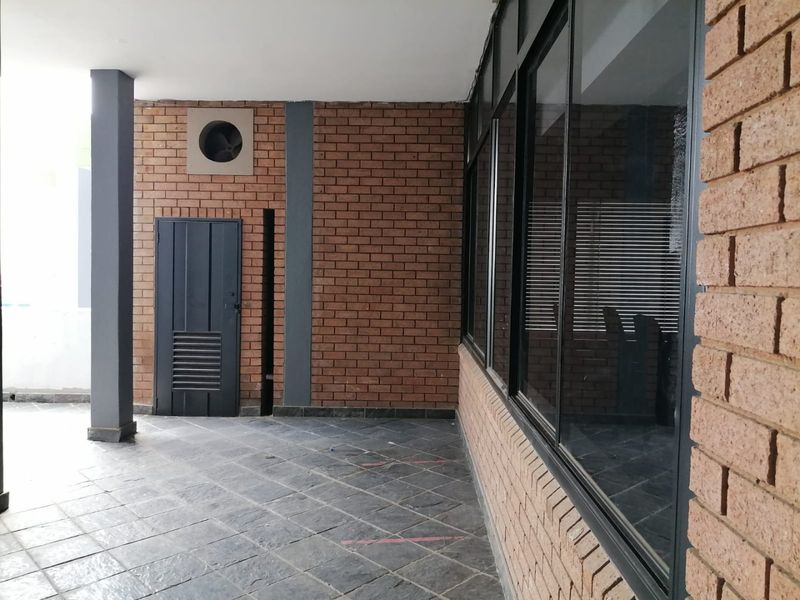 A covered outdoor patio area featuring exposed brick walls, tiled flooring, and large glass windows, providing a functional transition space for the commercial property.