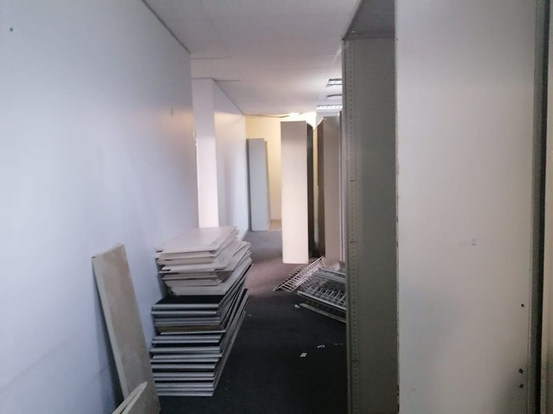 A narrow commercial hallway featuring a suspended ceiling and recessed lighting, currently in a state of disrepair with visible wall damage.
