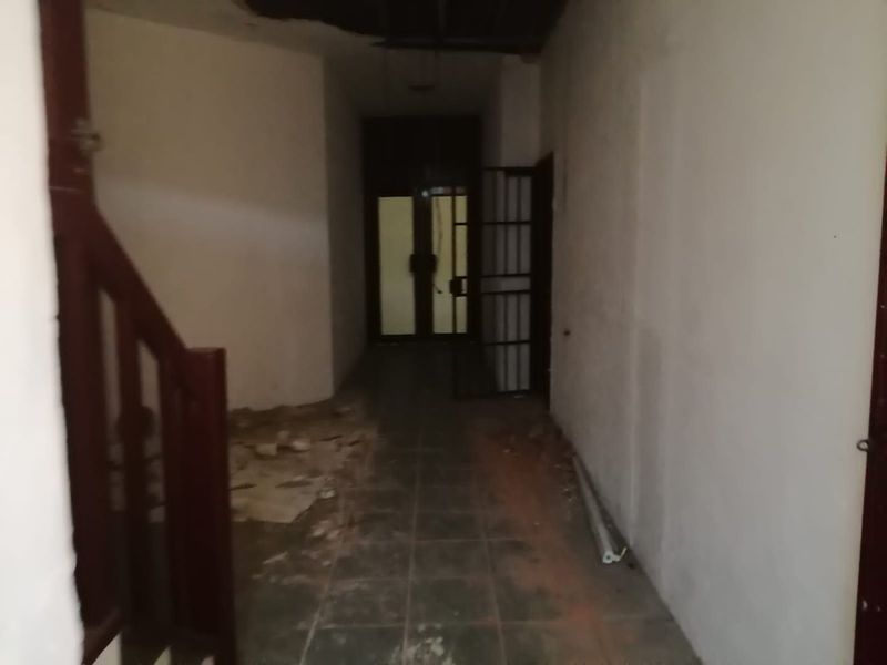 A vacant, dilapidated hallway featuring tiled flooring, a security gate, and visible ceiling damage, requiring significant renovation and repair.
