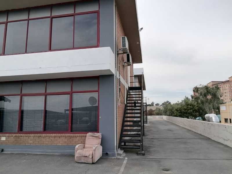 Rear exterior view of a commercial building featuring large windows, external metal staircase, and wall-mounted air conditioning units on a paved area.