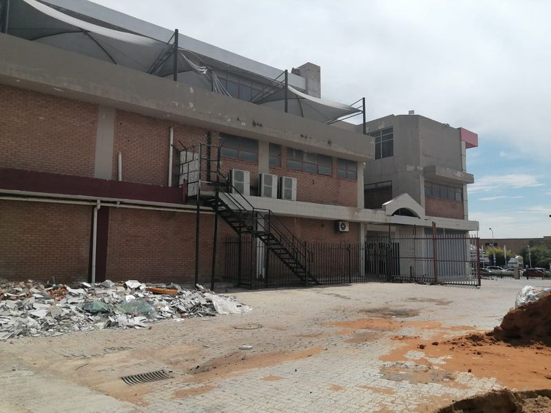 Rear exterior view of a commercial building featuring brick facade, metal staircase, security fencing, and a paved yard area with visible debris.