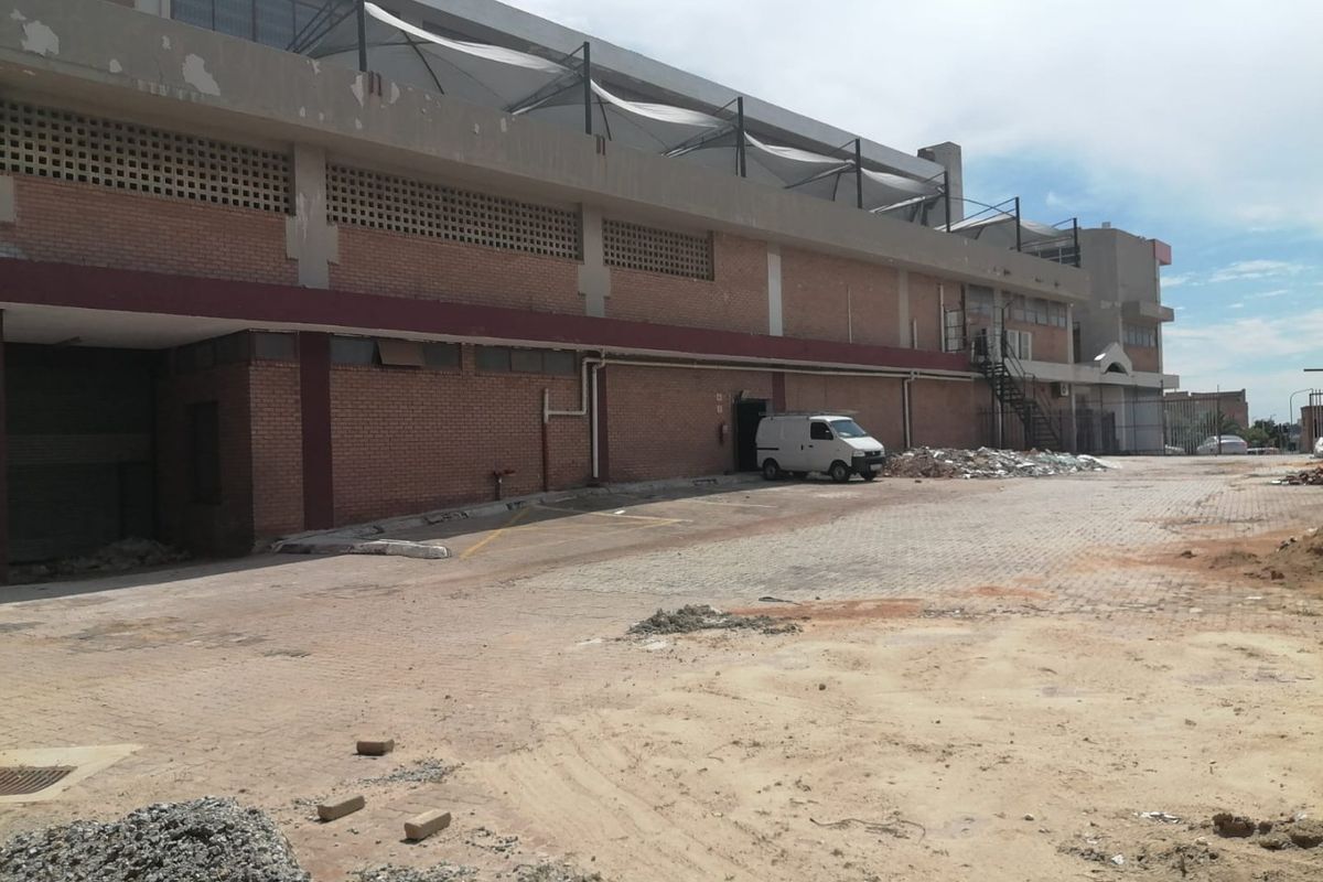 Rear exterior view of a commercial building featuring brick facade, roller shutter access, and a large paved yard area requiring maintenance.
