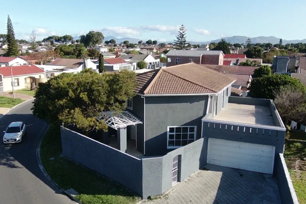 Aerial view of a detached house featuring a grey exterior, tiled roof, paved driveway, and a secure boundary wall in a suburban neighborhood.