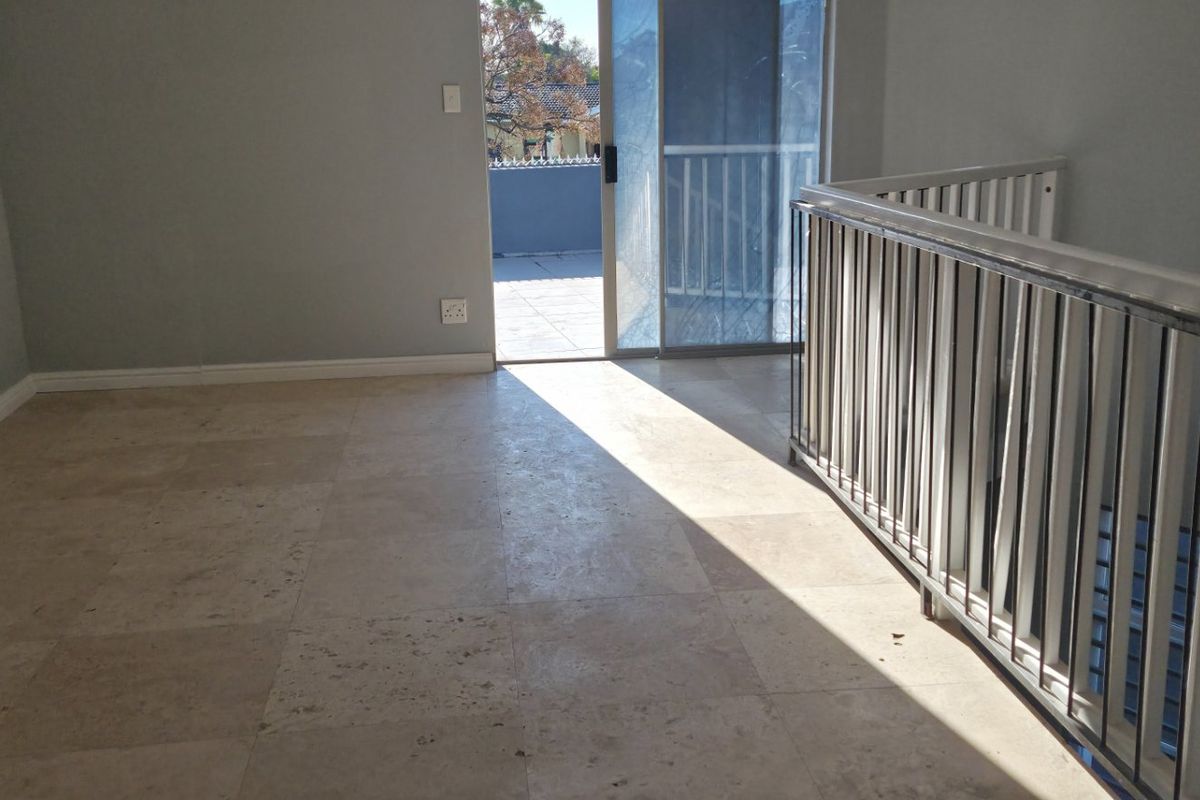 An empty living area featuring travertine flooring, a metal balustrade overlooking the staircase, and direct access to an outdoor balcony through a sliding door.