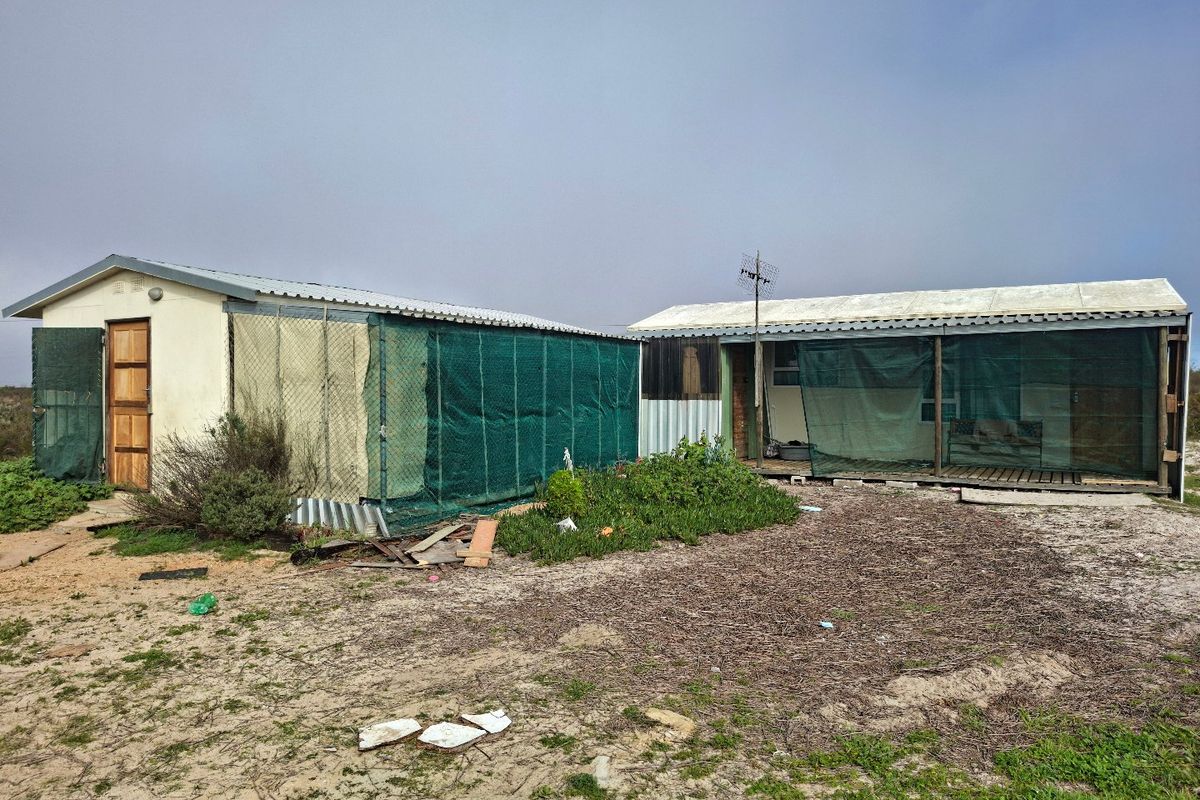 Rear view of a residential structure featuring corrugated metal roofing, timber decking, and exterior shade netting on a sandy plot.