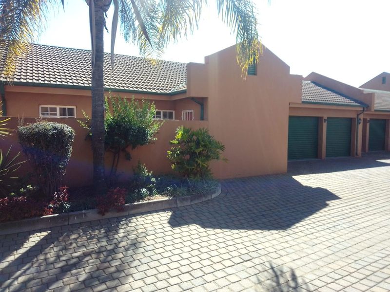 Front elevation of a townhouse featuring a paved driveway, tiled roof, and multiple roller shutter garage doors with established garden landscaping.