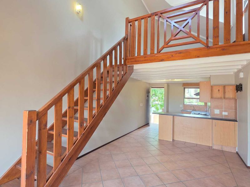Open-plan living area featuring a wooden staircase leading to a mezzanine level, tiled flooring, and a functional kitchen with built-in cabinetry.
