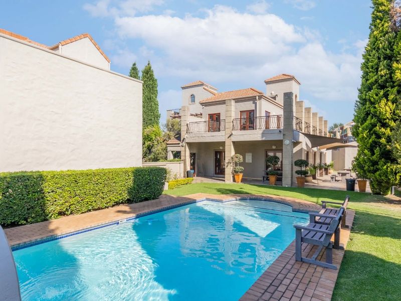 A well-maintained outdoor pool area featuring a swimming pool with brick coping, surrounded by a landscaped garden and a two-story building with a balcony.