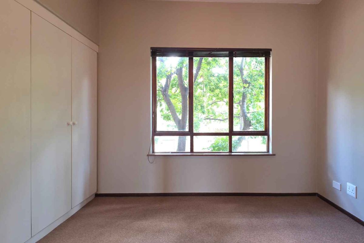 A bright, empty bedroom featuring neutral walls, carpet flooring, and built-in cupboards with a large window overlooking greenery.
