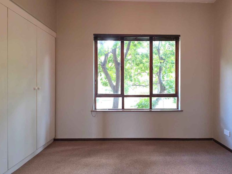 A bright, empty bedroom featuring neutral walls, carpet flooring, and built-in cupboards with a large window overlooking greenery.