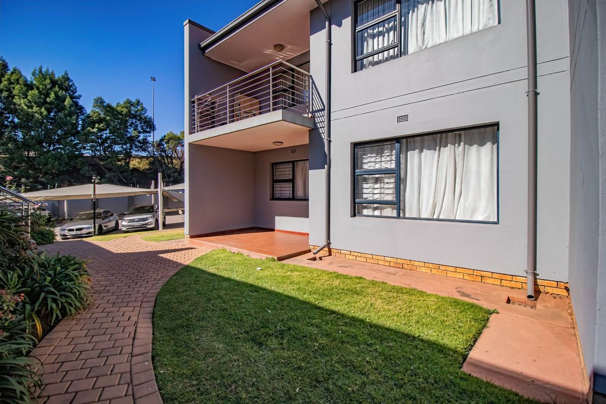 Rear view of the apartment building featuring a private balcony, paved garden area, and well-maintained exterior facade with aluminium window frames.