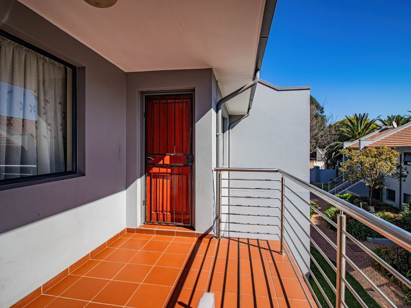 A clean, tiled balcony featuring a stainless steel balustrade and a secure entrance door, overlooking the complex grounds.