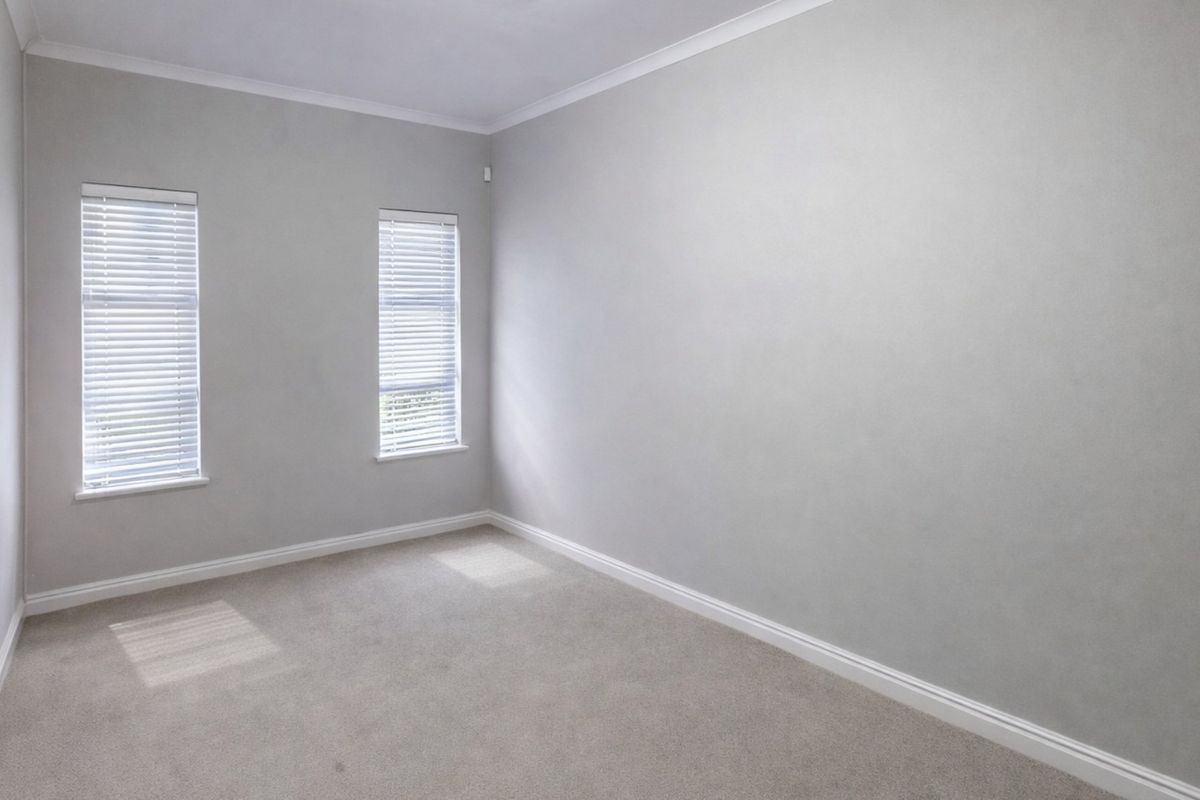 A clean, empty bedroom featuring neutral grey walls, carpeted flooring, and two windows fitted with venetian blinds.