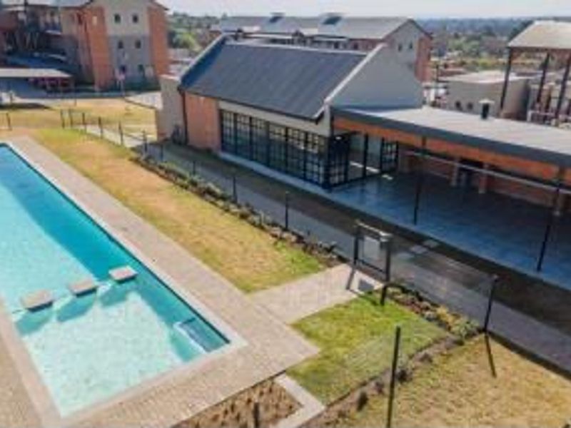 Aerial view of a communal swimming pool area featuring a paved deck, adjacent covered patio, and modern brick building architecture.
