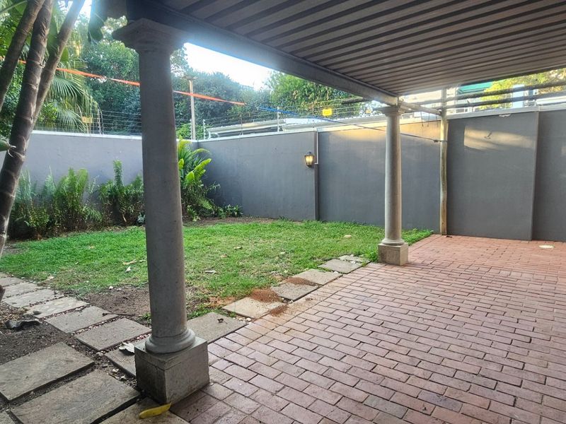 A covered patio area featuring brick paving and concrete columns, overlooking a private garden enclosed by perimeter walls with electric fencing.