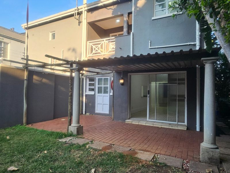Rear view of the property featuring a covered brick-paved patio, a small garden area, and an upper-level balcony.