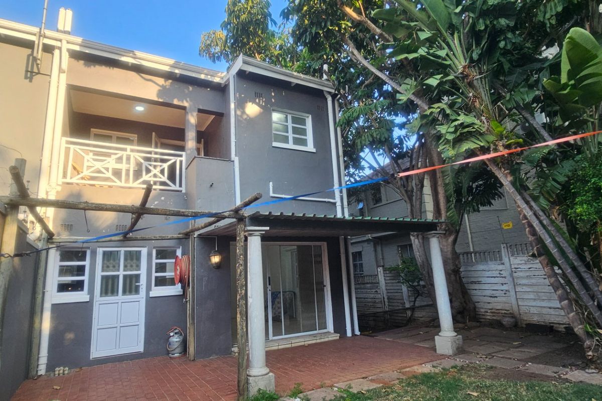 Rear view of a two-story house featuring a covered patio, balcony, and paved outdoor area with established garden surroundings.