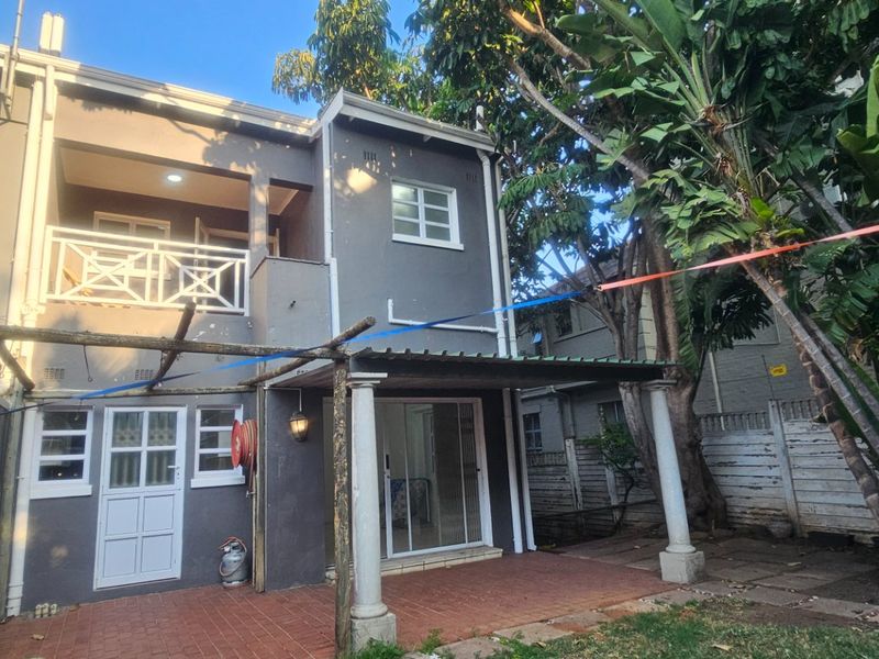 Rear view of a two-story house featuring a covered patio, balcony, and paved outdoor area with established garden surroundings.