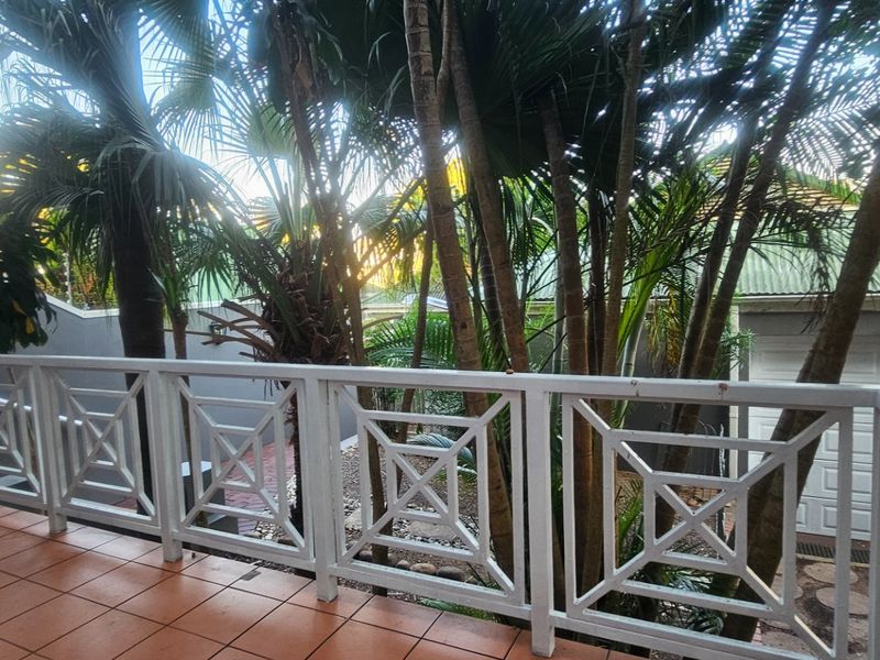 A covered patio area featuring tiled flooring and a decorative white balustrade, overlooking a lush garden and garage structure.