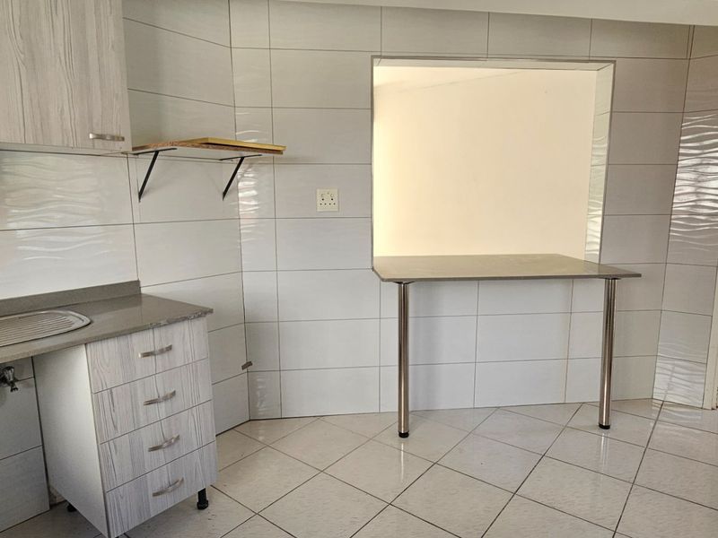 A functional kitchen area featuring tiled walls and flooring, built-in cabinetry, a stainless steel sink, and a breakfast bar opening into an adjacent room.