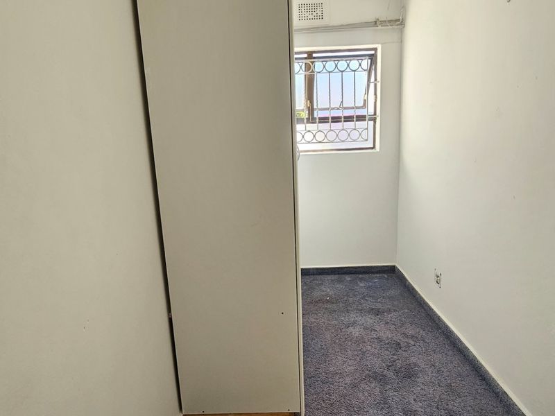A compact bedroom featuring carpeted flooring, a window with security bars, and a built-in cupboard, offering a functional space for a single bed or office.