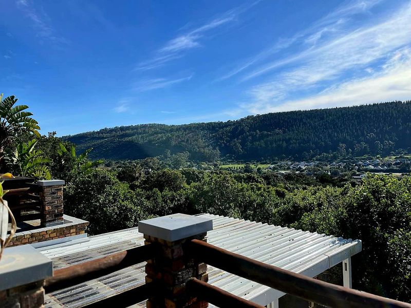 Elevated view overlooking a lush green valley and forested hills, featuring a foreground of a corrugated metal roof and brick-pillared balcony railing.