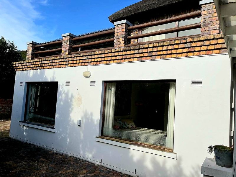 Rear exterior view of the house featuring a paved patio area, white rendered walls, and an upper-level balcony with brick detailing and thatched roofing.