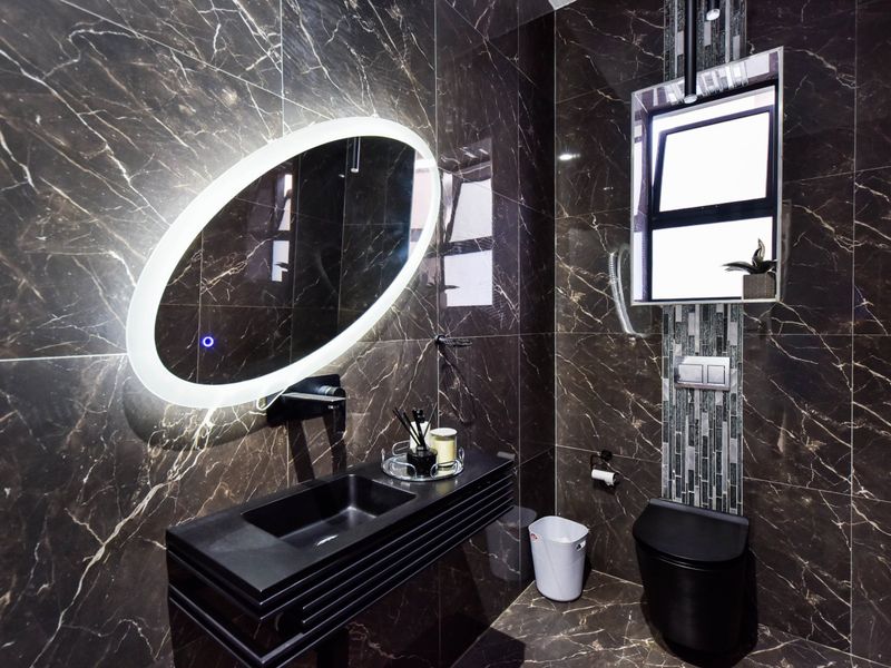 Modern bathroom featuring dark marble-effect tiling, a sleek wall-mounted vanity with integrated basin, a backlit oval mirror, and a wall-hung toilet.