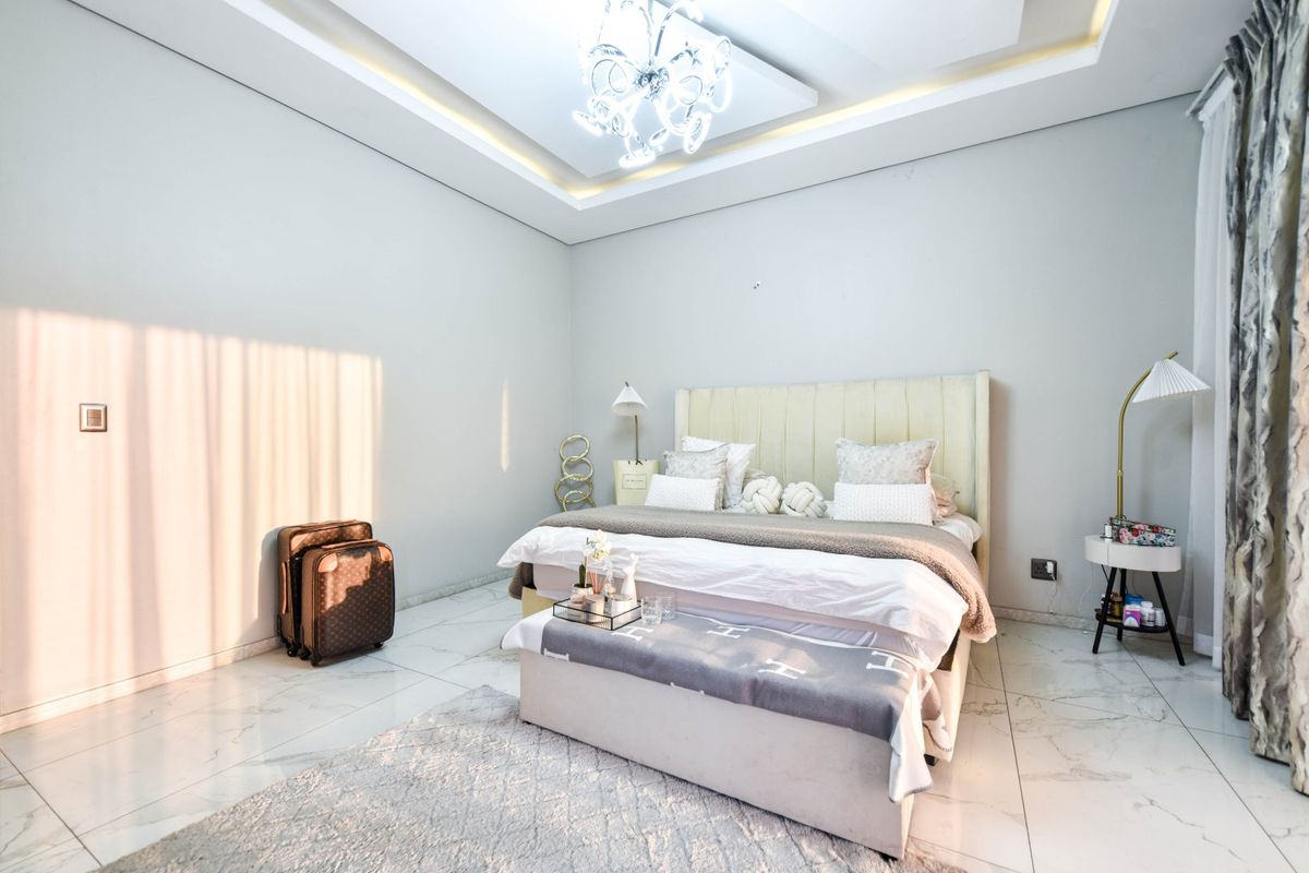 A well-maintained bedroom featuring marble-look floor tiles, a recessed ceiling with cove lighting, and a central chandelier.
