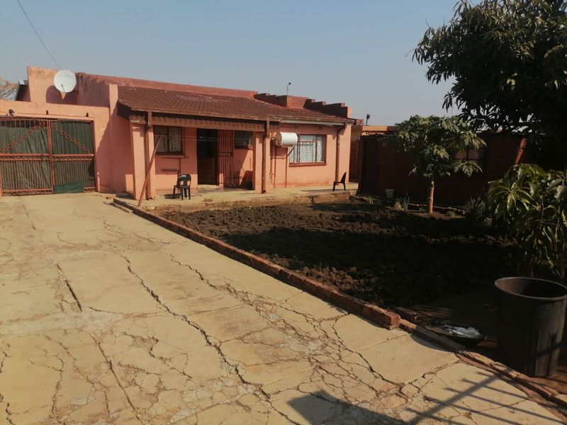 Front view of a single-story house featuring a tiled roof, metal security gate, and a large concrete driveway with visible surface cracking.
