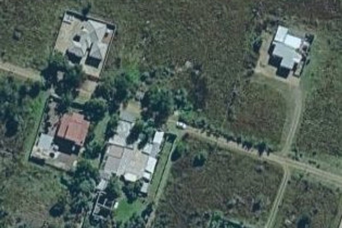 An aerial view of a residential area featuring multiple plots of land, surrounding vegetation, and unpaved access roads.