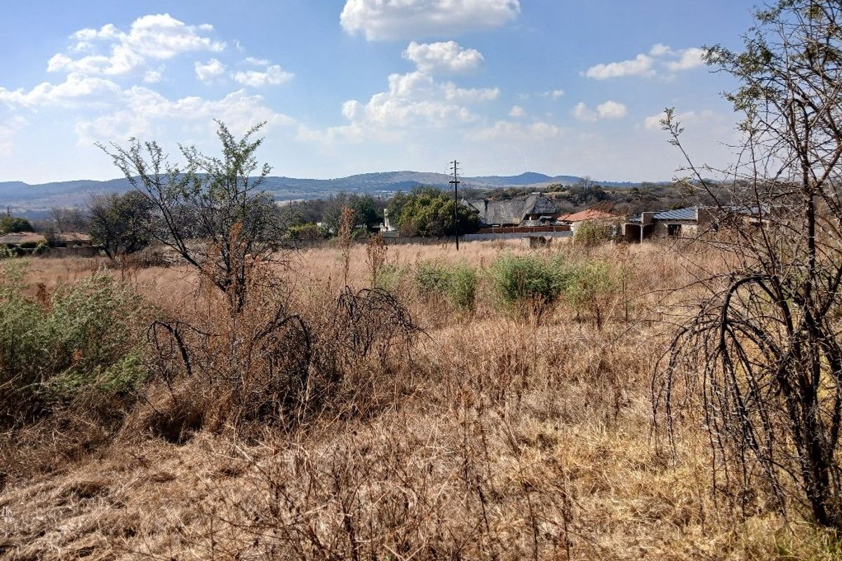 A view of a vacant residential plot featuring dry vegetation, surrounding boundary walls, and distant mountain views.